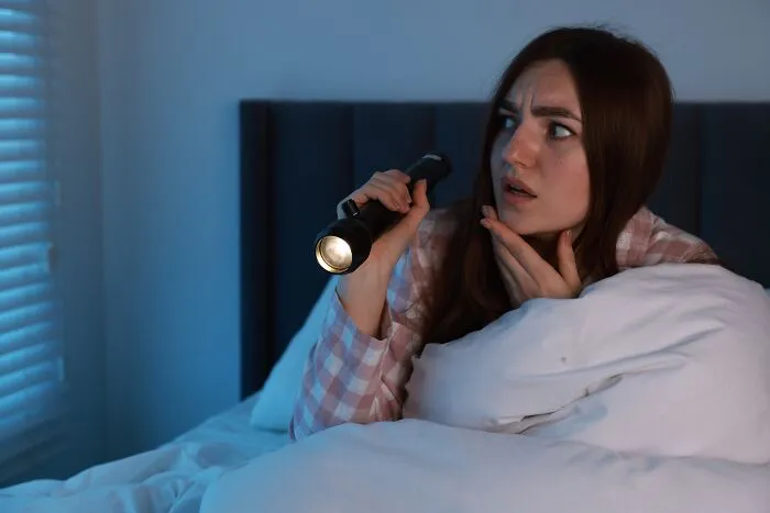 Scared woman in bed holding a flashlight, worried about spooky unexpected noises