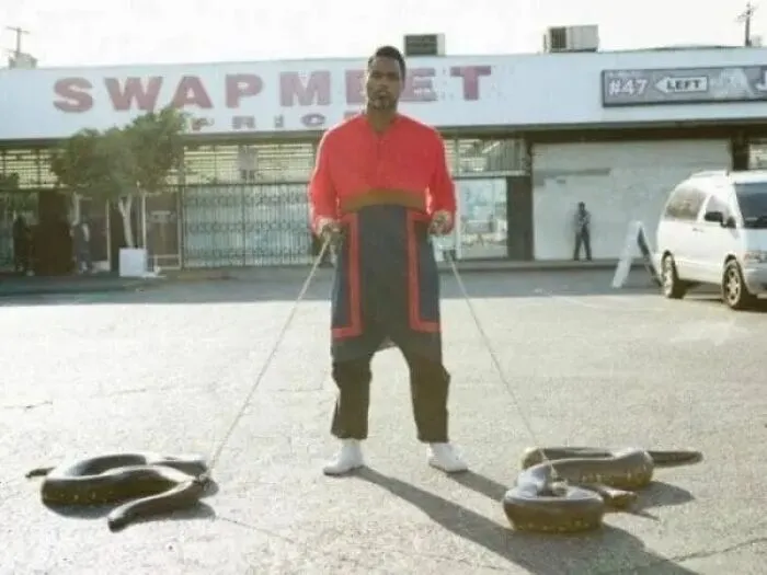 Man in bright clothing holding two large snakes on leashes in a parking lot