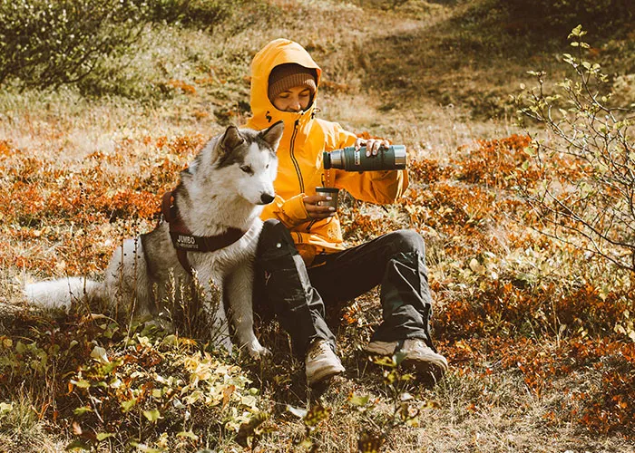 A person in a yellow jacket pouring a drink beside a husky dog outdoors, connected to online mysteries.