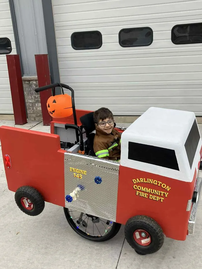 Smiling child in a firefighter costume inside a custom fire truck wheelchair frame for Halloween.