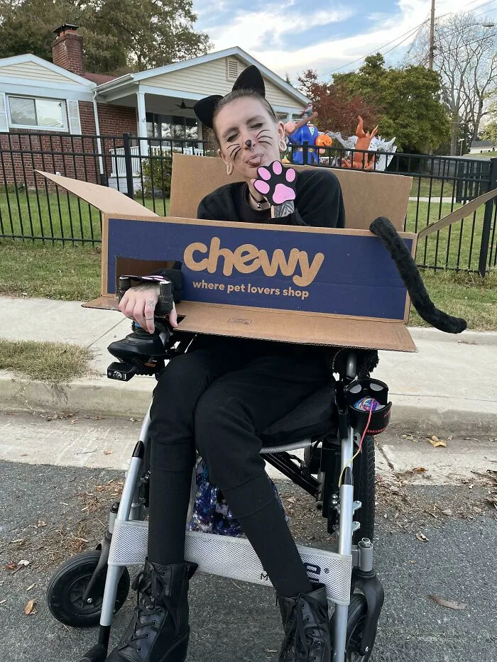 A person in a black cat costume sitting in a wheelchair styled like a cardboard Chewy box for Halloween.