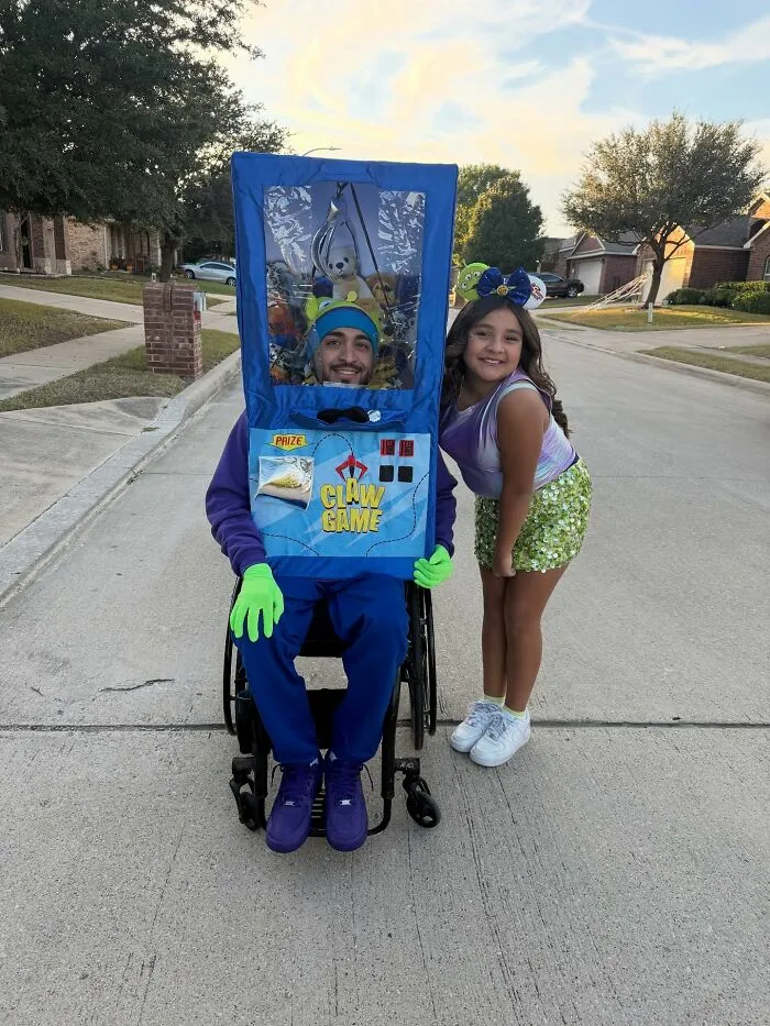 A man in a wheelchair formatted as a claw game machine poses with a girl for Halloween.
