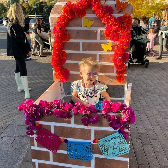 Little girl smiling inside a colorful Encanto themed floral booth wheelchair costume for Halloween.