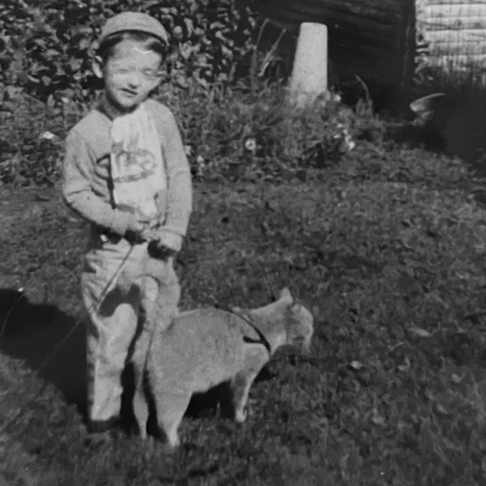 A vintage photo of a young boy holding a leash on a small cat outdoors