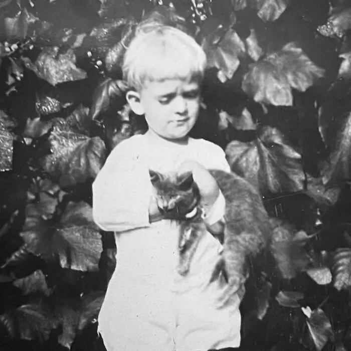A young child holding a small cat in front of leaves in vintage photos of children with their cats