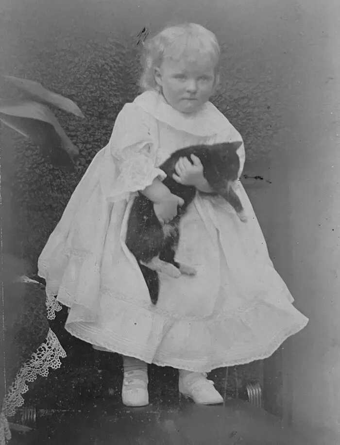 A young child in a ruffled vintage dress carefully cradling a dark kitten in photos of children with their cats