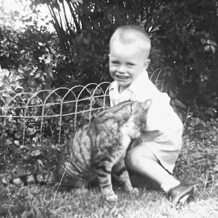 A smiling young boy kneeling in a garden next to a striped tabby cat in vintage photos of children with their cats