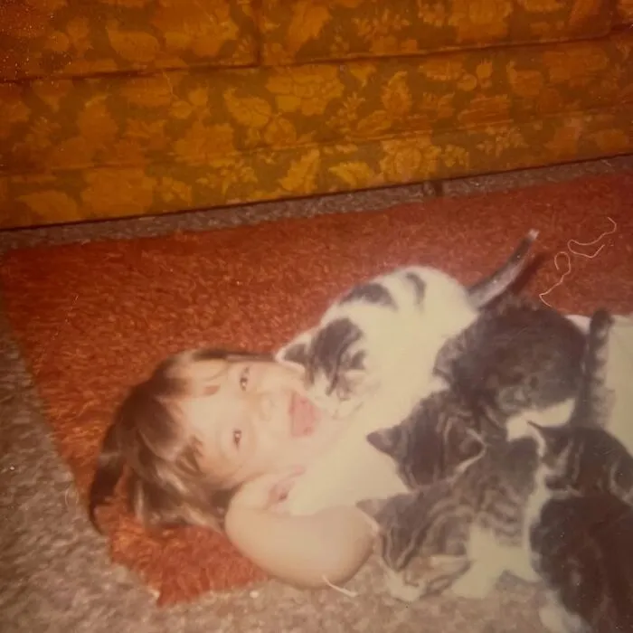 A child laughs while lying down covered in kittens in vintage photos of children with their cats