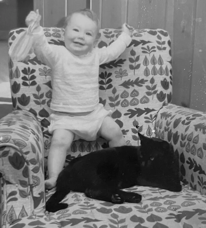 A happy toddler sits in an armchair with a black cat from these vintage photos of children with their cats