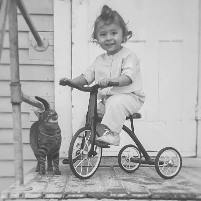 A happy toddler riding a tricycle next to a pet cat, from vintage photos of children with their cats