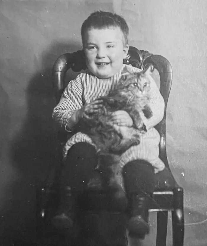 Smiling child sitting in a wooden chair holding a fluffy cat, from vintage photos of children with their cats