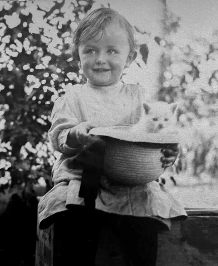 A classic vintage photo of a smiling young child holding a kitten inside a hat