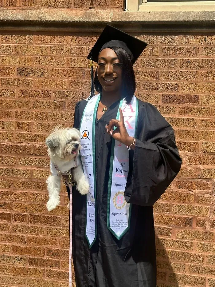 A happy graduate in cap and gown holding a small white dog, sharing life stories with her pet.