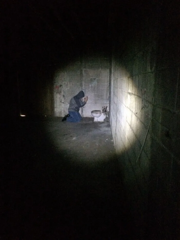 Person kneeling by broken toilet in dark room lit by flashlight beam