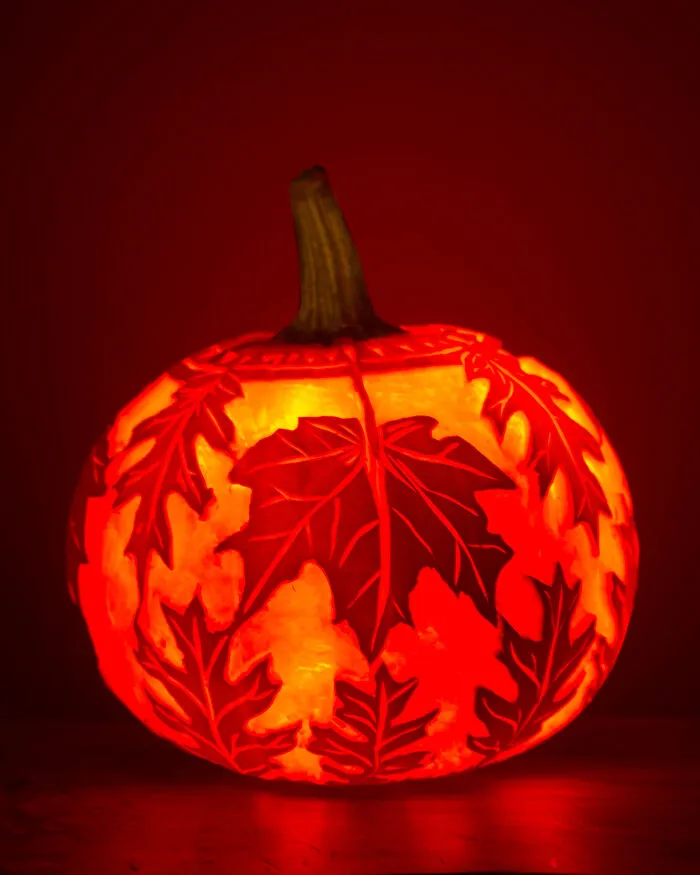 A glowing Halloween jack-o-lantern pumpkin intricately carved with detailed leaf patterns.