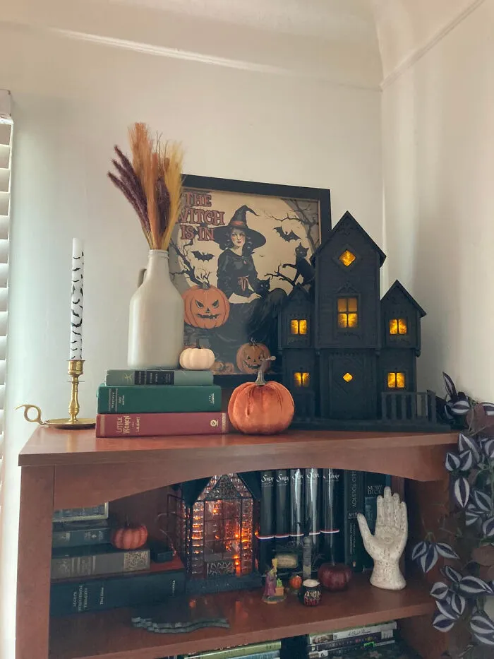 A bookshelf decorated for Halloween with a miniature haunted house, pumpkins, books, and spooky art.