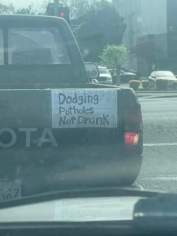 A pickup tailgate with a paper sign reading Dodging Potholes Not Drunk as a funny moving joke by truckers.