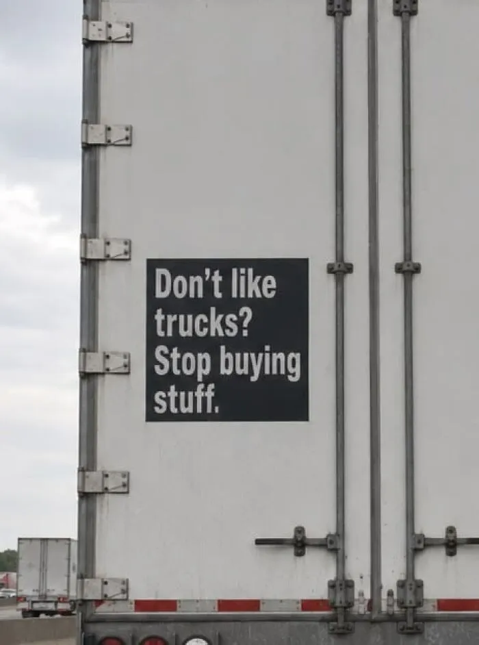 Funny sign on a semi-truck back door reading Don't like trucks? Stop buying stuff.