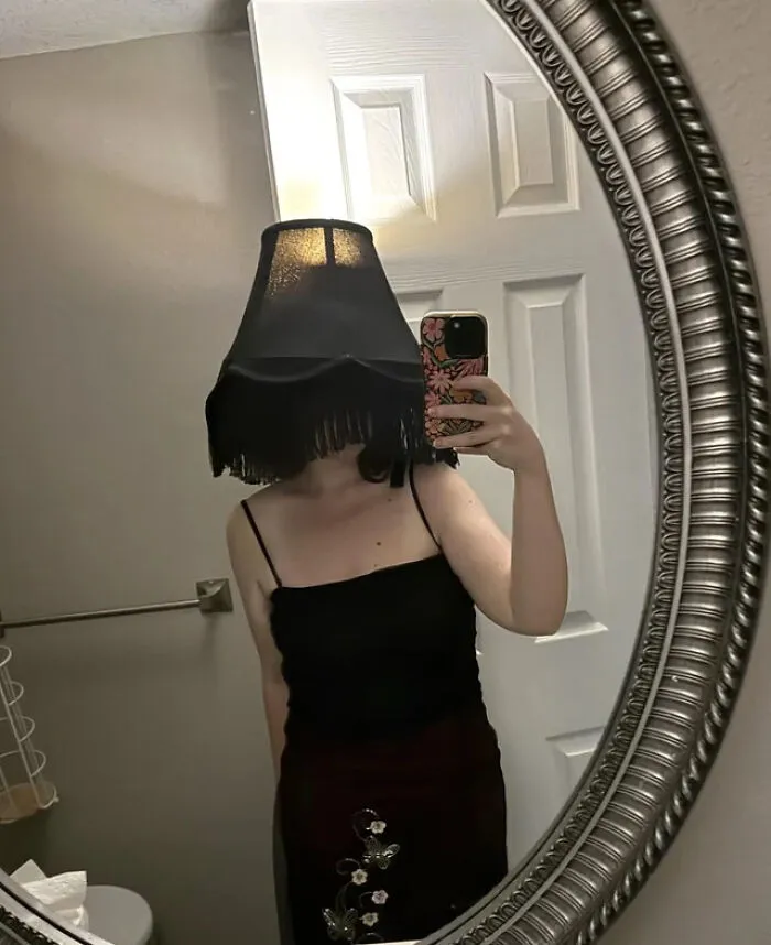 A woman taking a mirror selfie wearing a lampshade on her head for a cool Halloween costume idea.