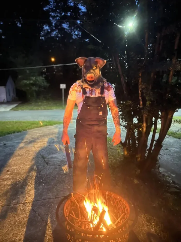 Person in a scary pig mask and overalls standing by a fire, showing creepy Halloween costume ideas.