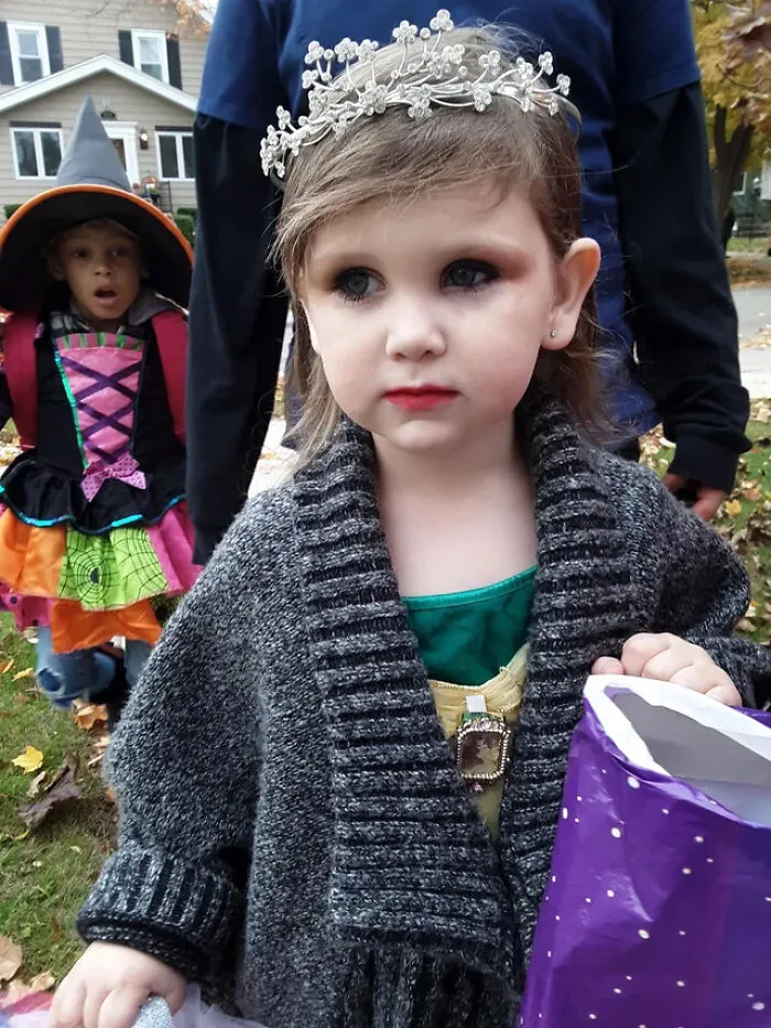 A young girl dressed up in a princess tiara and sweater for a cute Halloween costume while holding a trick-or-treat bag.