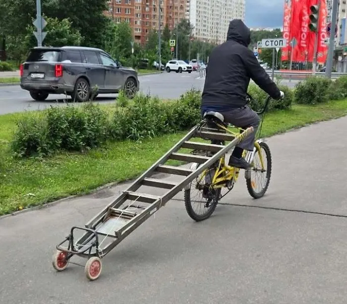A cyclist towing a ladder on small wheels, demonstrating resourceful Slavic engineering on the street.
