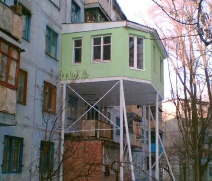 A green balcony extension built on long metal supports showing Slavic engineering on an apartment building.
