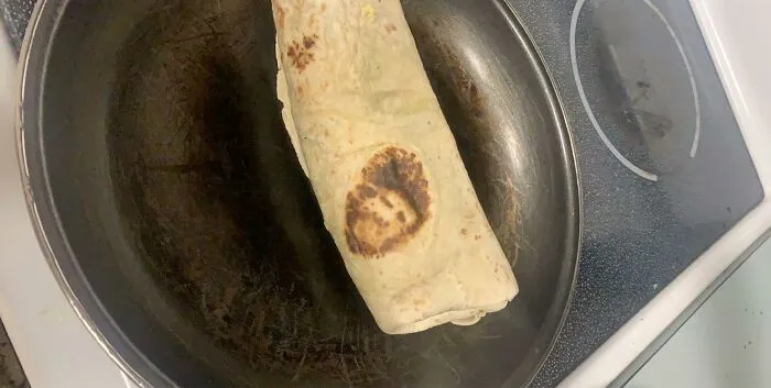 A perfectly confusing photo of a grilled burrito in a pan with a toasted spot resembling a human face.