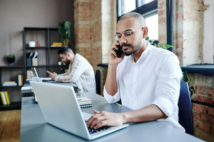 A man working on a laptop while talking on the phone in an office, reading wild mil stories.