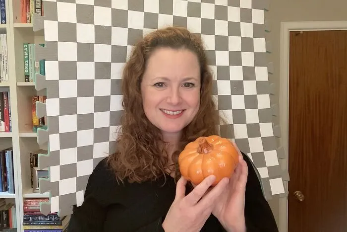 Woman holding an orange pumpkin in front of a checkerboard pattern for creative Halloween costume ideas.