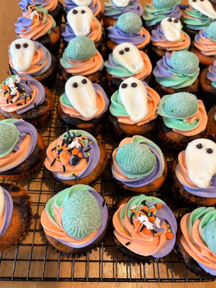 Colorful Halloween foods like cupcakes decorated with frosting ghosts and brain shapes on a wire rack.