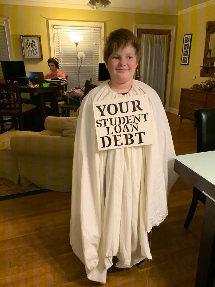 A kid dressed as a ghost with a sign reading Your Student Loan Debt for funny Halloween costumes.