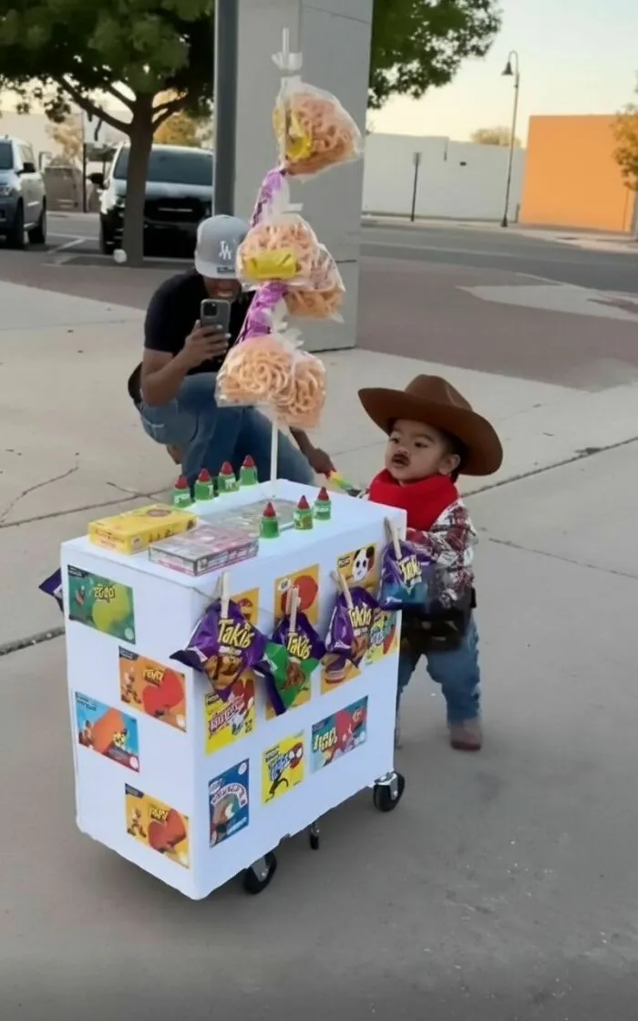 A toddler wearing a snack vendor Halloween costume pushing a white cart filled with chips and candy.