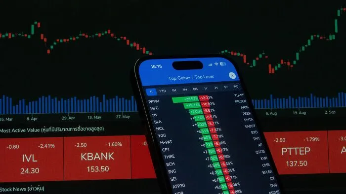 Smartphone showing a stock market trading app next to financial charts, illustrating money-saving tricks.