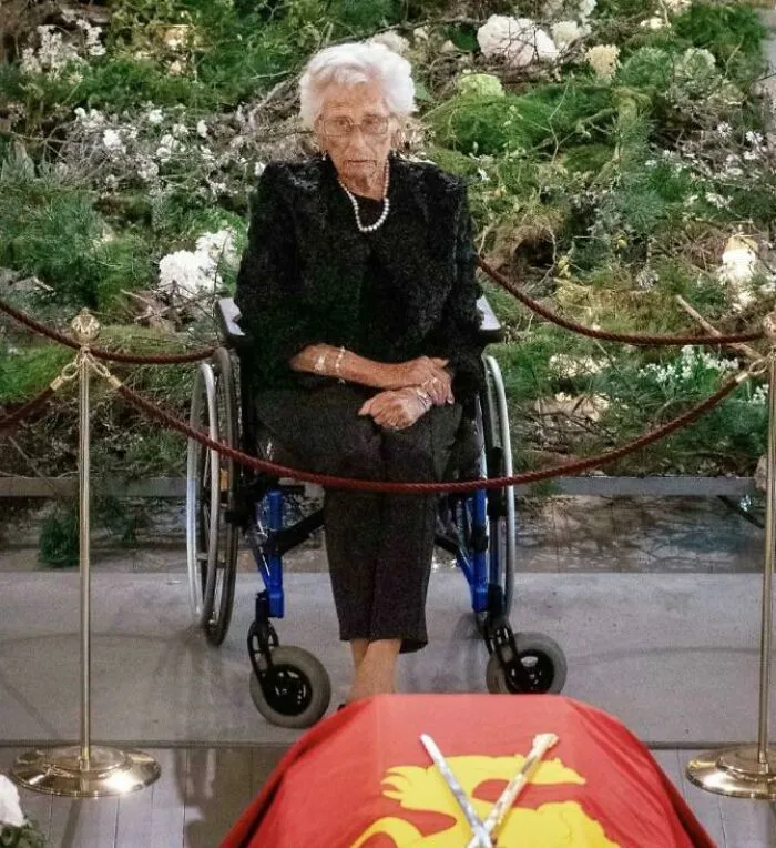 An elderly woman sitting in a wheelchair near a red casket with flowers captured in photos.
