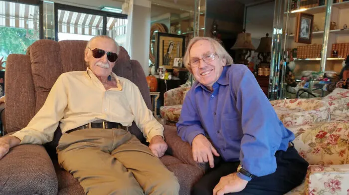 Stan Lee sitting in a armchair smiling with another man, among people captured in photos for the last time.