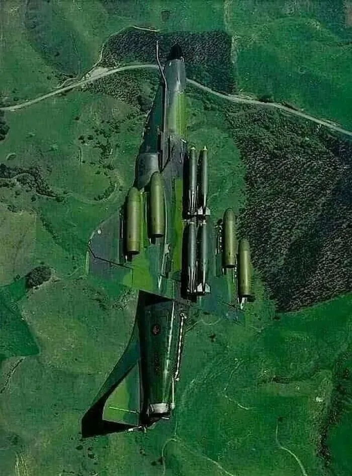 Camouflaged fighter jet flying over green fields, an impressive optical illusion photo.