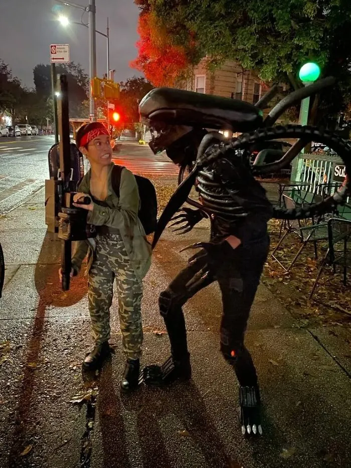 Funny Halloween couples costume featuring Ripley and a Xenomorph alien standing on a city sidewalk at night