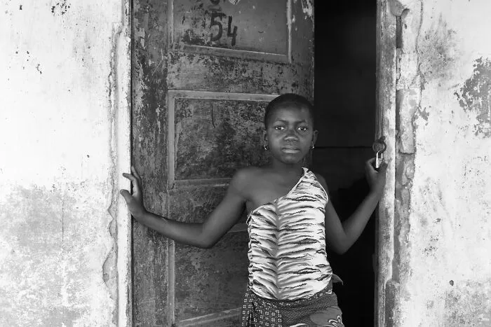 A black and white portrait photo of a young girl standing in a rustic doorway looking ahead