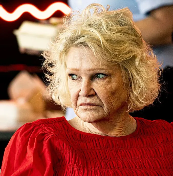 A close-up portrait photo of an elderly blonde woman with a serious expression in a red shirt