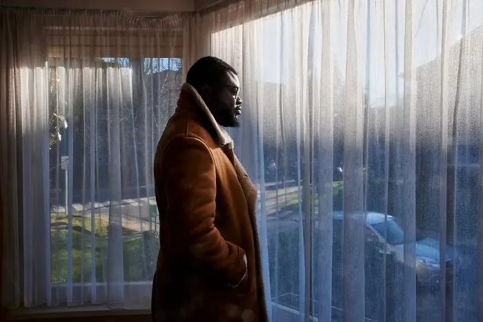 Moody portrait photos of a man in a brown coat looking out a window behind sheer curtains