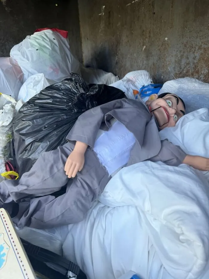 A weird and creepy ventriloquist dummy lying on top of trash bags in a dumpster.