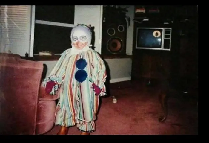 A child standing in a dimly lit living room wearing a weird and creepy clown costume.