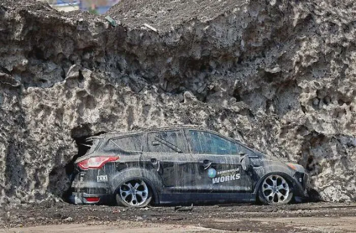 A dirty car parked under a massive wall of melted snow in weird photos.