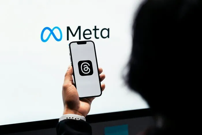 Hand holding smartphone displaying Threads app icon in front of Meta logo on computer screen, representing top tech companies