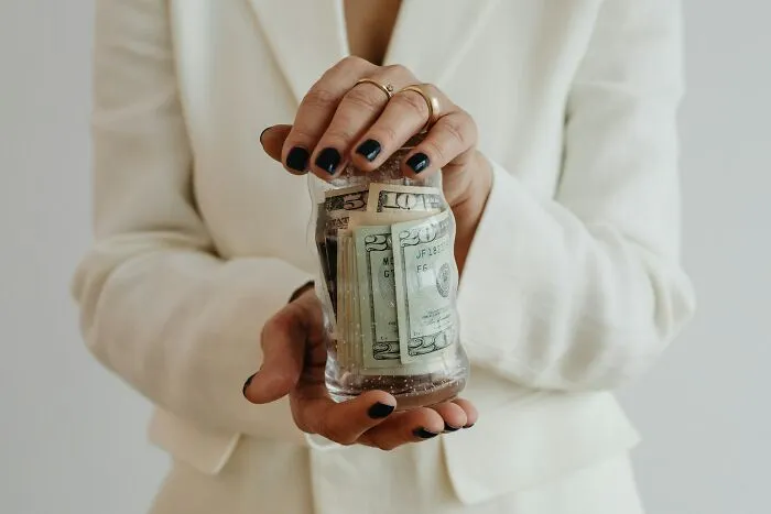 A person in a white blazer holding a glass jar filled with US dollar bills, offering brilliant tips on saving money