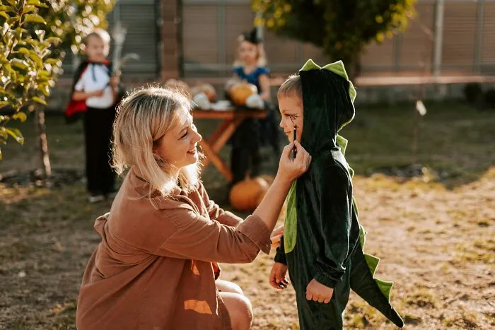 Woman applying face paint to a child wearing a green dinosaur costume for Halloween
