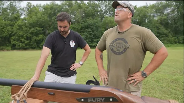 Two men standing outdoors beside a huge prop gun in a perfectly confusing perspective.