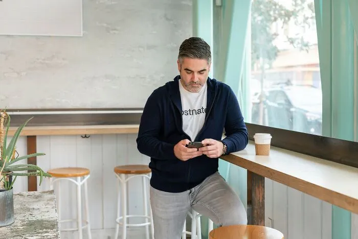 A middle-aged man standing near a window looking at his phone, representing adult children of narcissistic parents