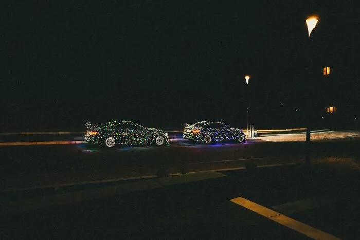 Two sports cars illuminated with colorful Christmas lights at night, creating a Hollywood-style scene.